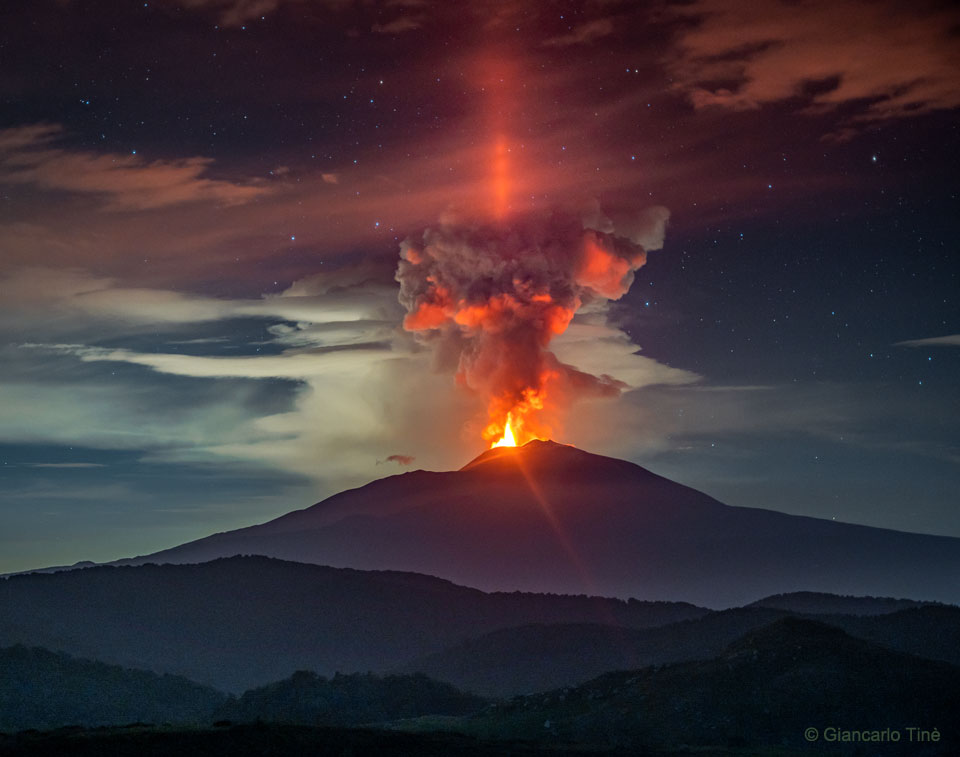 Zdjęcie ukazuje czerwony filar światła, widoczny nocą pod Etną, wybuchającym wulkanem we Włoszech. 
Bardziej szczegółowe informacje w opisie poniżej.