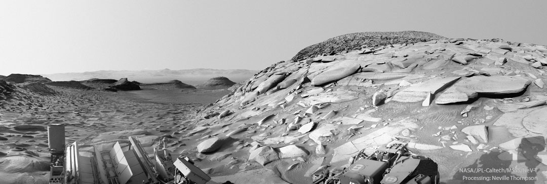 A view of Mars from the Curiosity rover on Mars 
is pictured in black and white. Many rocks and hills are 
visible, with a hill containing many unusually flat rocks 
visible on the right.
Please see the explanation for more detailed information.
