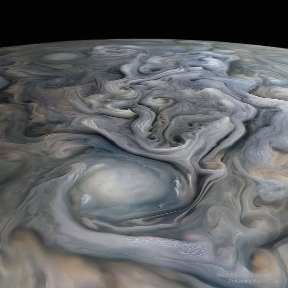 The cloud tops of Jupiter are pictured in a closeup
flyby of the Juno spacecraft. A big white oval cloud is
visible in the foreground, while many swirls of many muted
colors are visible trailing behind. A dark night sky is
in the background. 
Please see the explanation for more detailed information.