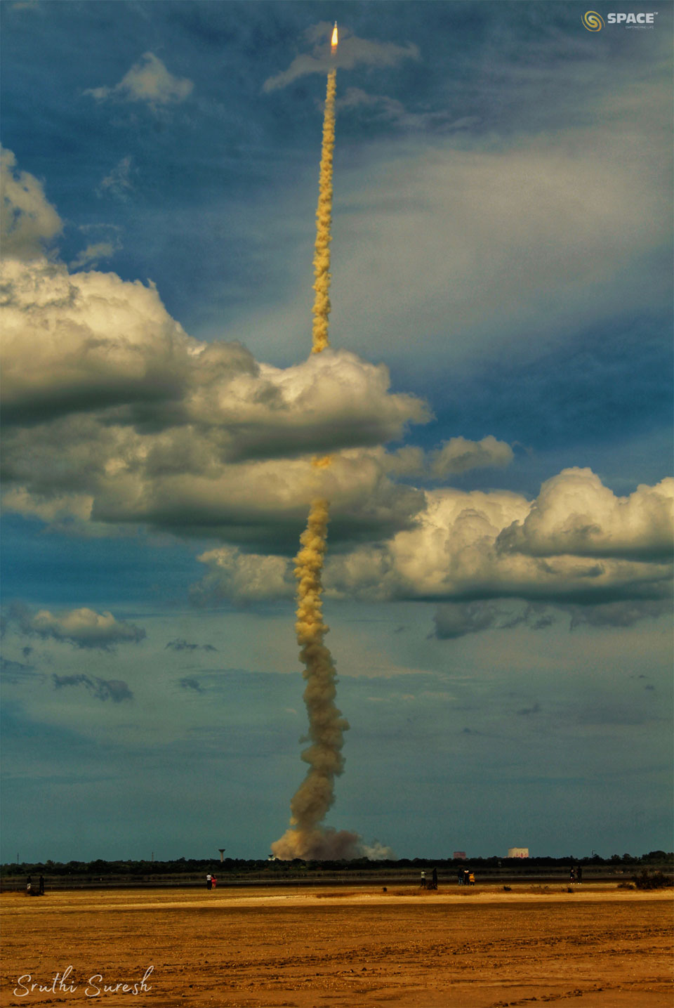 A rocket is seen after lift-off with a long smoke plume.
The rocket is captured against a blue sky and has gone through
a cloud deck. In the foreground is an empty tan-colored field. 
Więcej szczegółowych informacji w opisie poniżej.