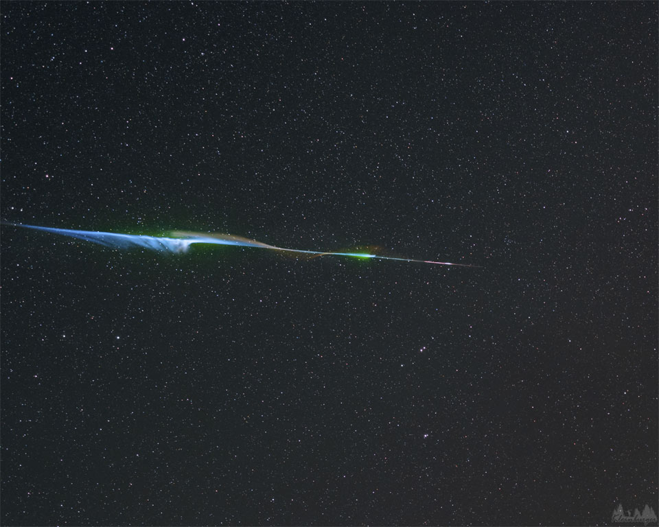 Centrum zdjęcia przecina jasny i barwny ślad meteoru, z którego
wypływają kosmki gazu. W tle widoczne jest pole gwiazd.
Zobacz opis. Po najechaniu kursorem myszki pokaże się opisana wersja,
po kliknięciu obrazka załaduje się wersja o największej dostępnej rozdzielczości.