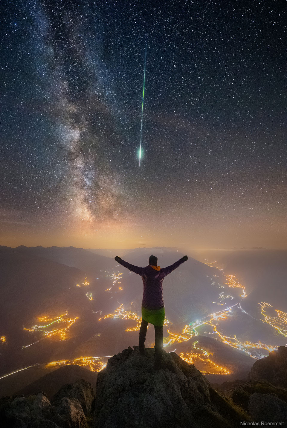 A person is seen facing away, standing on a peak. Other mountain peaks
surround them. City lights are seen in towns and along roads below. Stars
in the night sky are above. The band of the Milky Way galaxy slants down
from the upper left. A bright green meteor streak slants down from above.
Więcej szczegółowych informacji w opisie poniżej.