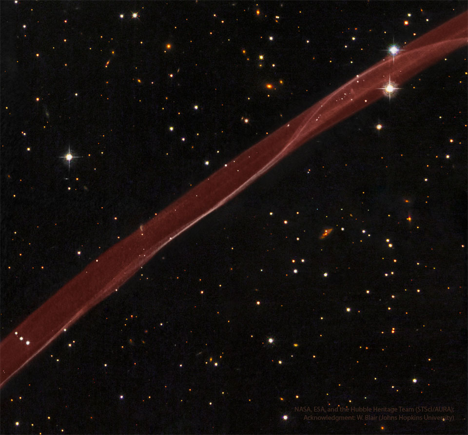 A thick transparent ribbon of red gas runs from the lower left to
the upper right. A dark starfield with stars and galaxies surrounds the 
bright red ribbon.
Więcej szczegółowych informacji w opisie poniżej.