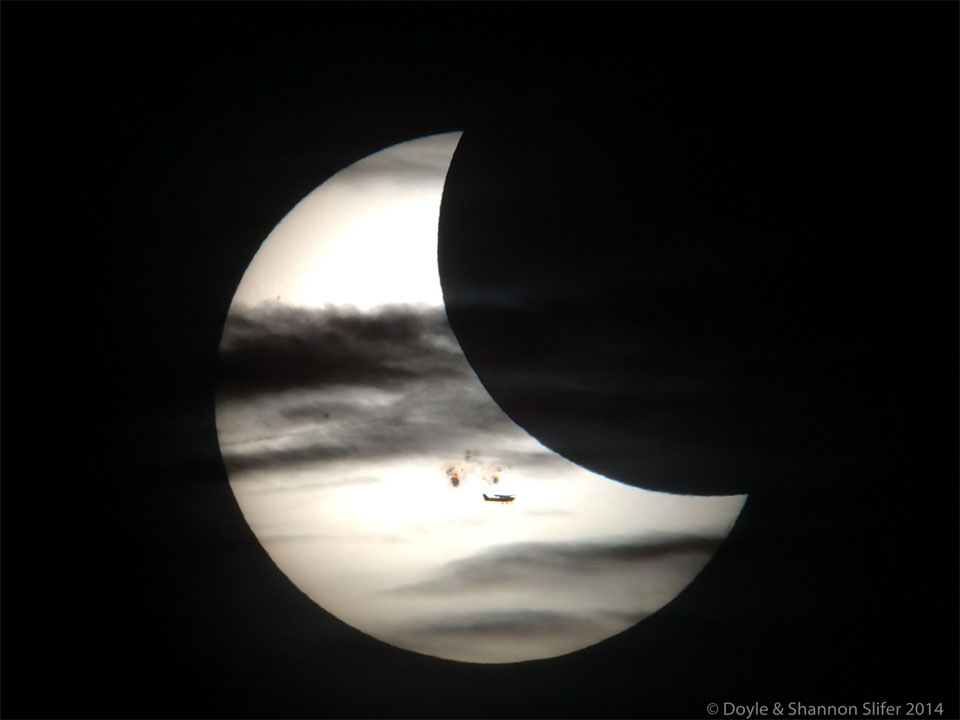 A partially eclipse Sun is shown. In front of the Sun 
are sunspots, the Moon, clouds, and an airplane.
Więcej szczegółowych informacji w opisie poniżej.