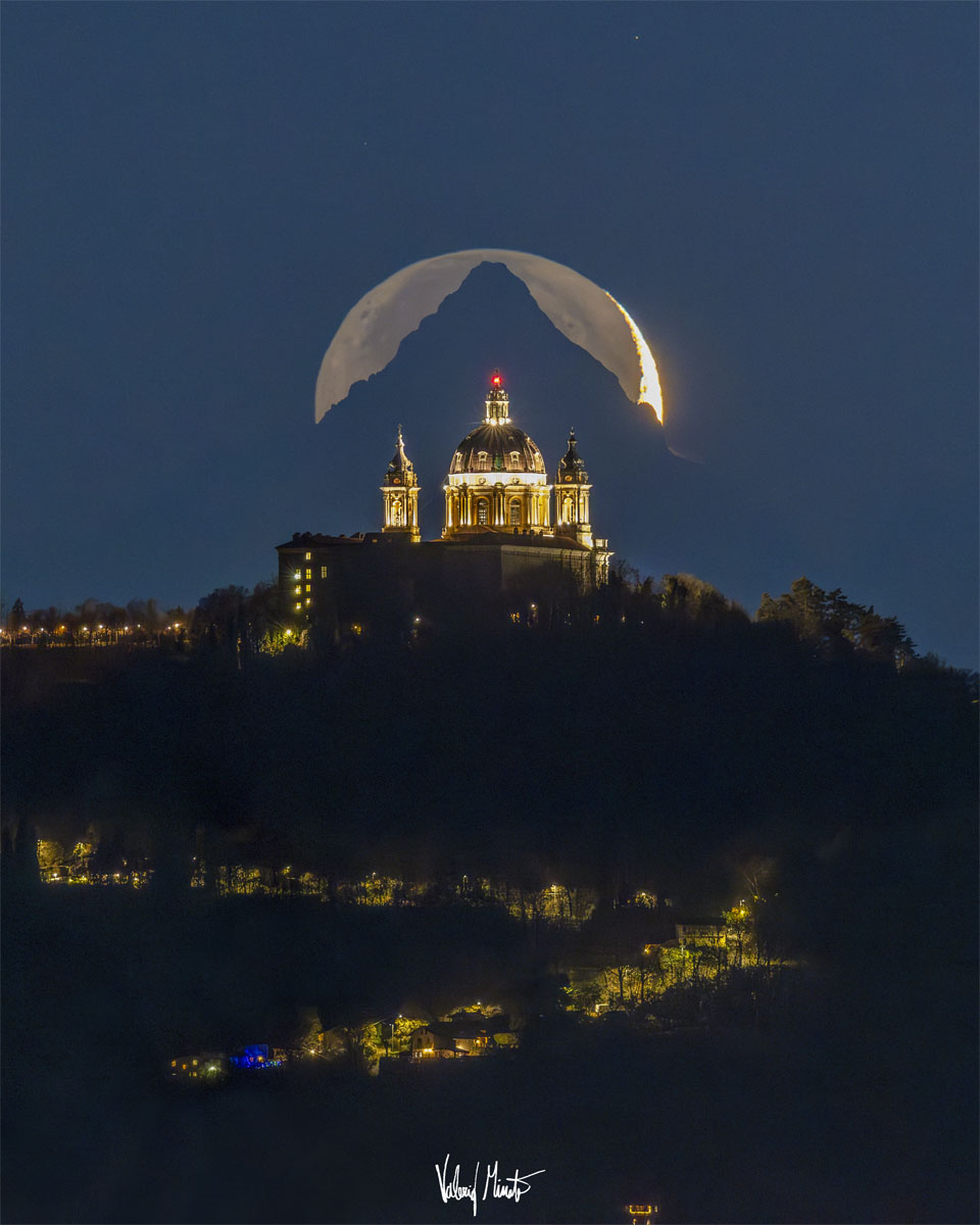 A tree-lined hill is shown topped by a majestic
cathedral. Directly behind the cathedral is 
of a triangular-shaped mountain top. Directly behind
the mountain is a crescent moon, although the exposure
is long enough to see the rest of lunar circle. 
Więcej szczegółowych informacji w opisie poniżej.