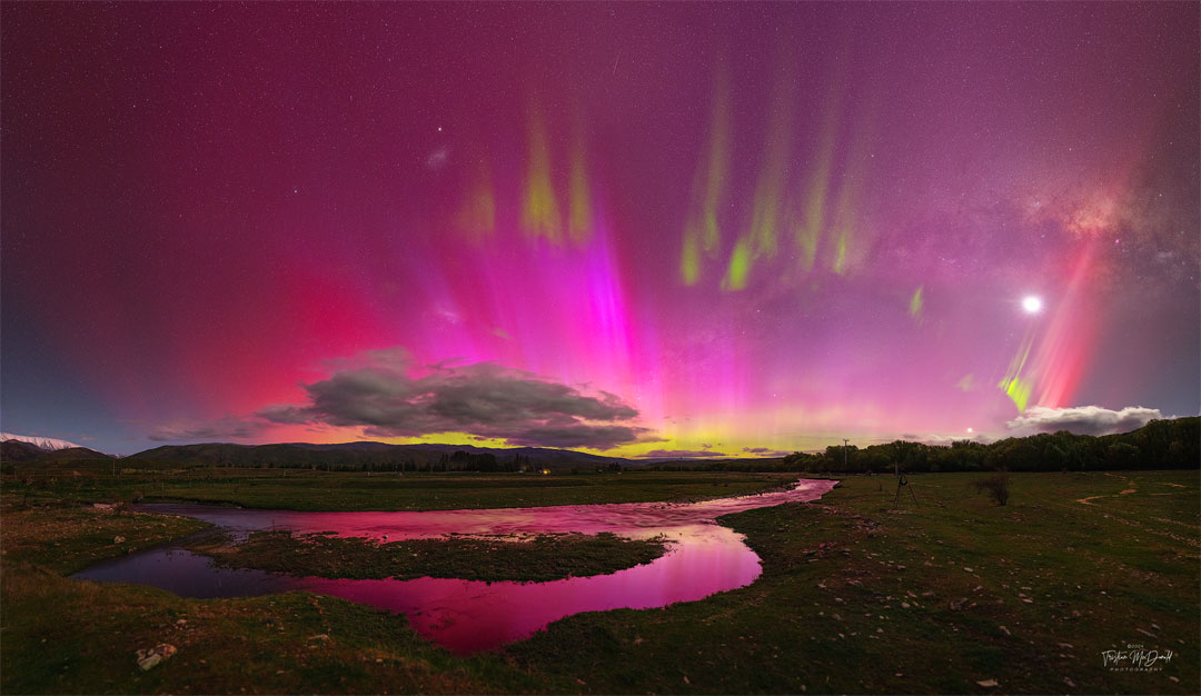 A night sky is shown that appears mostly red due to pervasive
aurora. In the foreground is covered by watery grasslands. Clouds 
are visible above the horizon. Thin green aurora are visible 
toward the top of the frame. In the background one can find the Moon,
the LMC, SMC, Venus, a meteor, and the band of our Milky Way galaxy.
Więcej szczegółowych informacji w opisie poniżej.