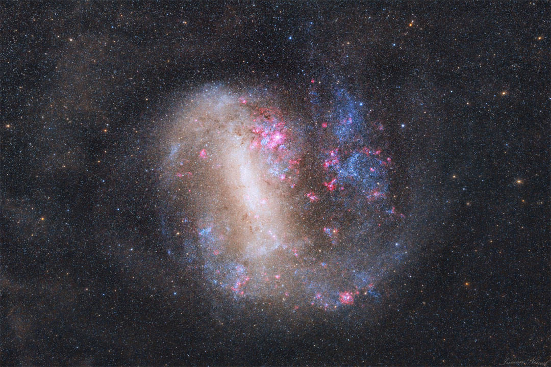 An unusual looking galaxy is shown with a light bar
running nearly vertical and blue stars and red nebulas 
around the edges.  
Więcej szczegółowych informacji w opisie poniżej.