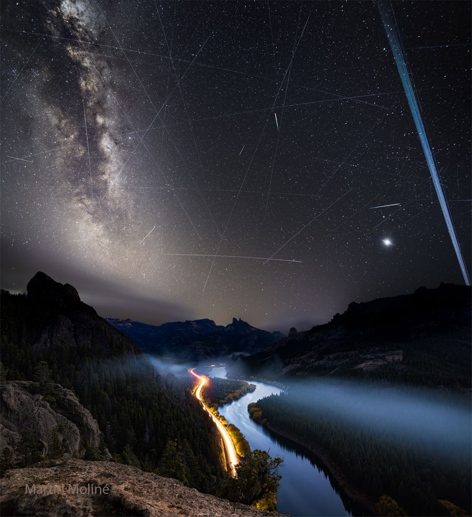 A night sky is shown with many stars and streaks.
In the foreground at the bottom are hills, a river, and
the red and white streaks of car lights.
Więcej szczegółowych informacji w opisie poniżej.