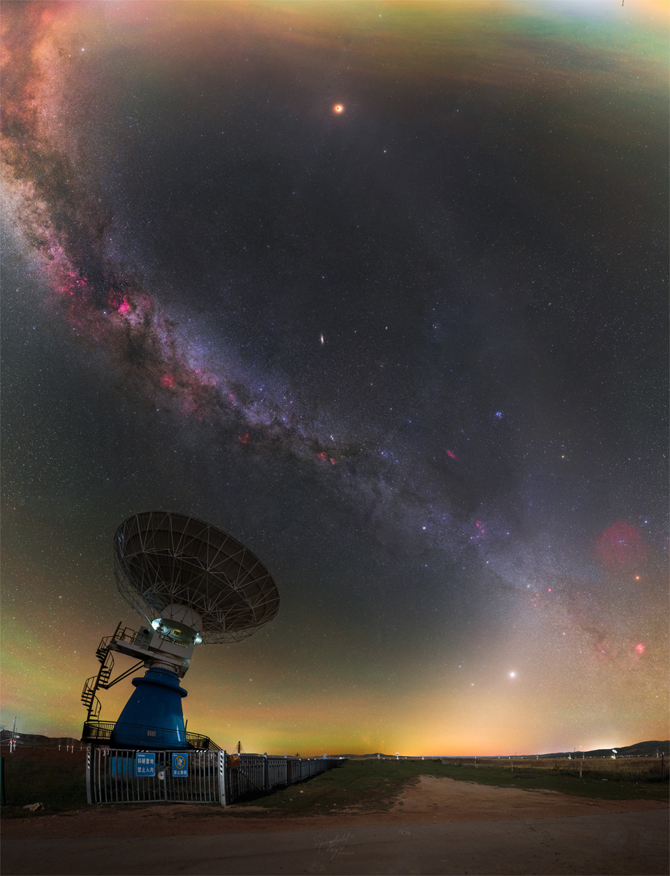 A wide angle image of the night sky is punctuated by
a radio telescope on the lower left. Two bands of light
run through the sky: the Milky Way and zodiacal light.
The bands intertwine like a double helix. Near the top
is an orange orb that is a totally eclipsed moon.
Więcej szczegółowych informacji w opisie poniżej.