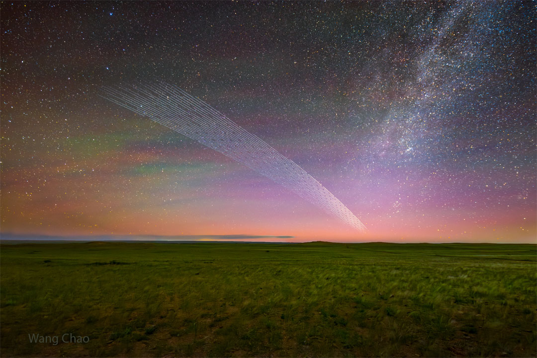 A star filled sky shows bands of green and purple sky glow. In
the foreground is a grassy field with clouds on the horizon. Most 
remarkably, a series of short streaks appear like a comet's
tail up from the horizon toward the upper left. 
Więcej szczegółowych informacji w opisie poniżej.