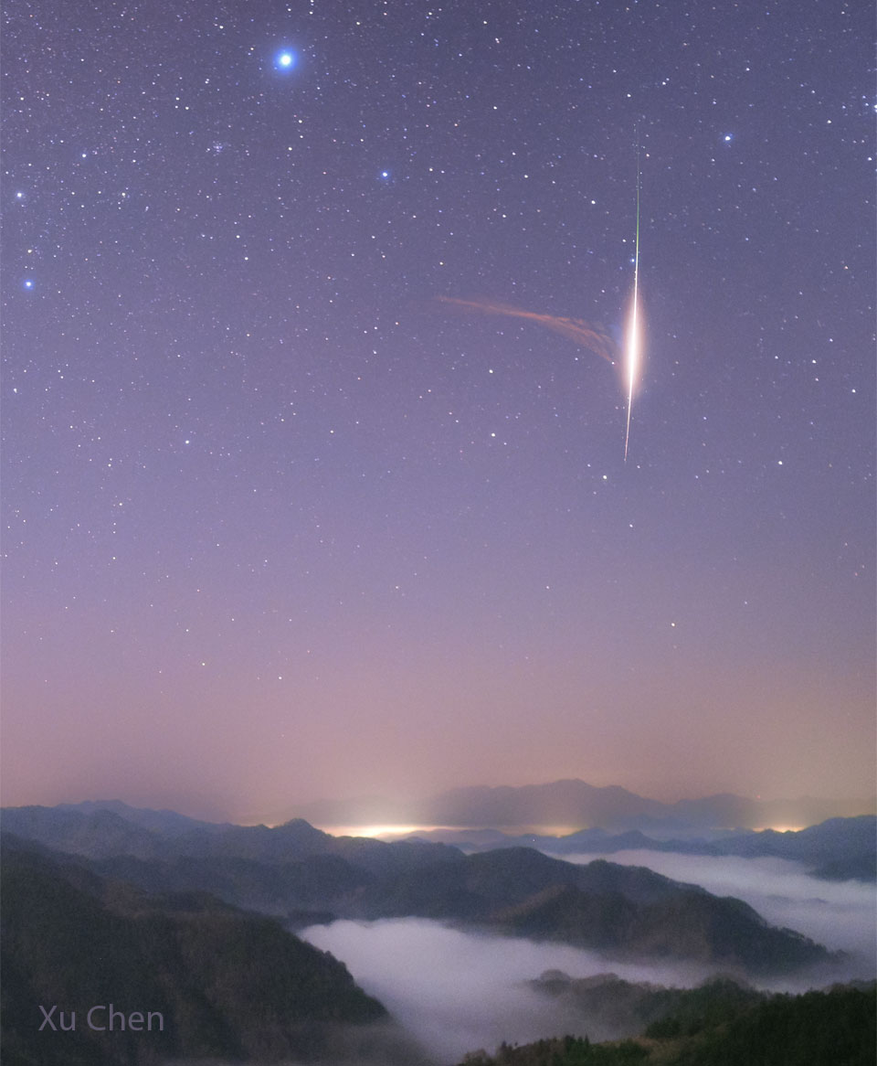A view of mountains over clouds shows a starfield
with a purple glow. Prominent on the right is the trail
of a bright meteor. To the left of the meteor and 
connecting to the meteor is something unusual: a light
brown triangular puff. 
Więcej szczegółowych informacji w opisie poniżej.