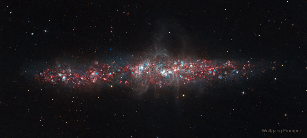 A galaxy is shown that has a lot of blue and red dots in it. The 
galaxy is an long elliptical blob across the middle and not a classic spiral. 
Więcej szczegółowych informacji w opisie poniżej.