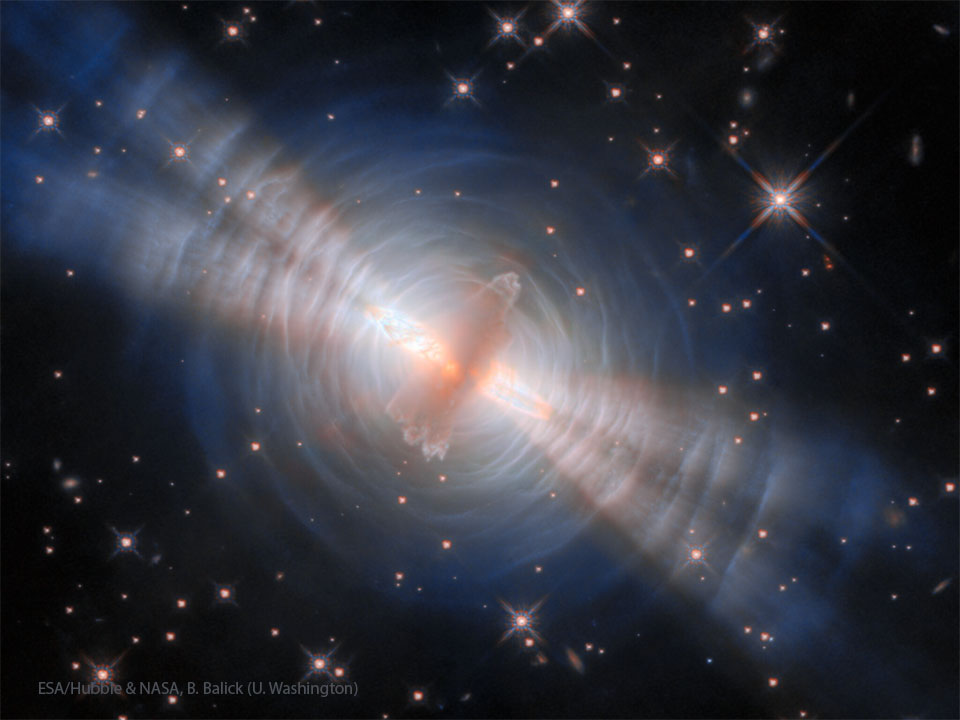 A starfield is shown with an unusual orange object in the center.
Surrounding this object are blue rings and four thick jets. 
Więcej szczegółowych informacji w opisie poniżej.