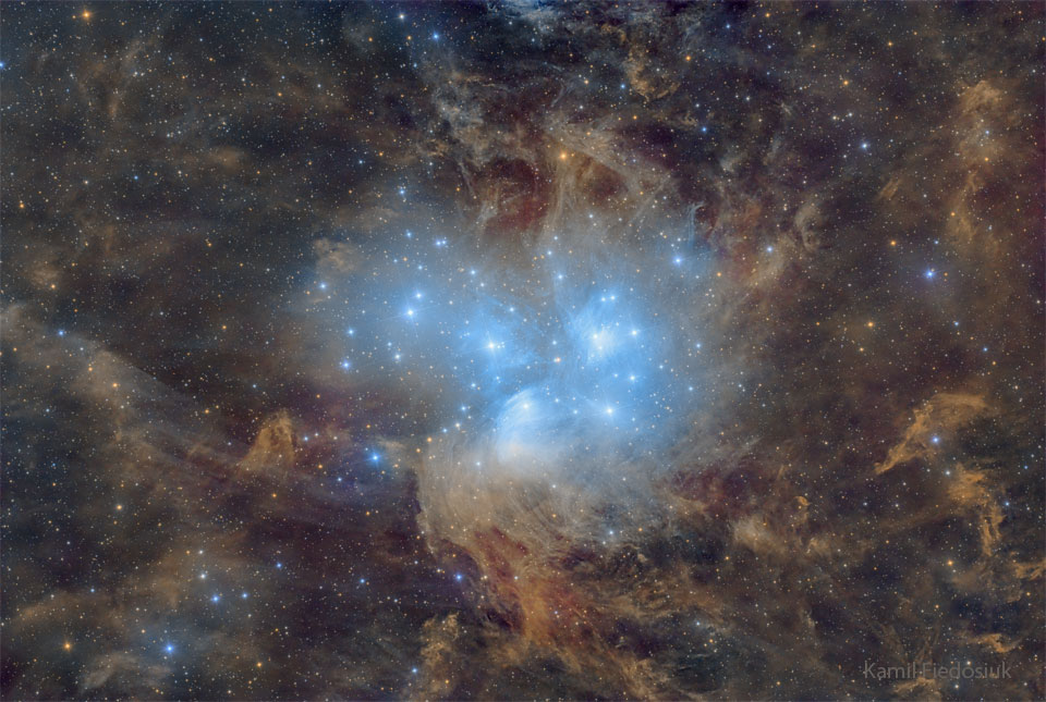 A cluster of blue stars is seen against a starfield
of brown and clumpy dust. The stars illuminate some of 
the nearby dust which causes the dust to glow blue.
Więcej szczegółowych informacji w opisie poniżej.