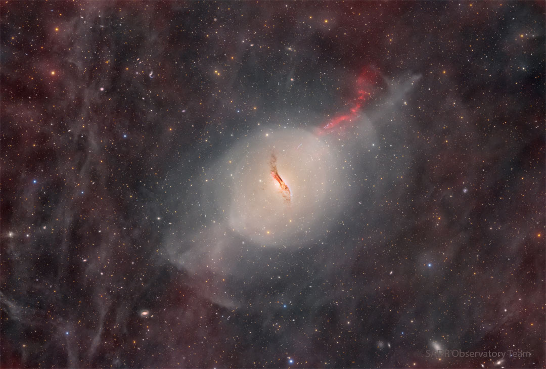 A starfield glows faintly with red glowing gas and dust.
In the center there is some sort of unusual galaxy with a red jet
emanating toward the upper right and a dark dust lane through
its center.  
Więcej szczegółowych informacji w opisie poniżej.