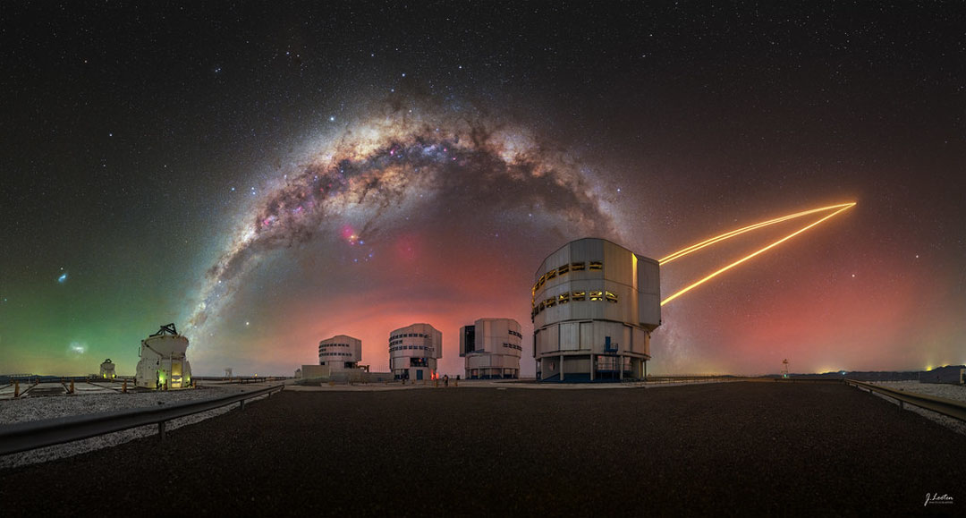 A colorful sky appears above several large telescopes.
Prominent in the sky is the band of the Milky Way Galaxy which
arches across the frame. Orange laser beams connect one
of the telescopes to the sky on the image right. 
Więcej szczegółowych informacji w opisie poniżej.