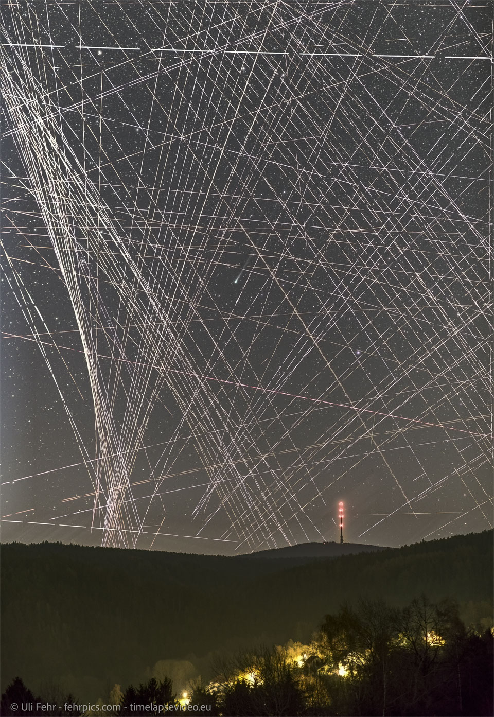 A night sky filled with lines and dashed-lines 
appears above a hilly landscape featuring a distant
lit-up tower. Near the center of the image is a comet
shown by its small green coma and ion tail to the upper 
right.
Więcej szczegółowych informacji w opisie poniżej.