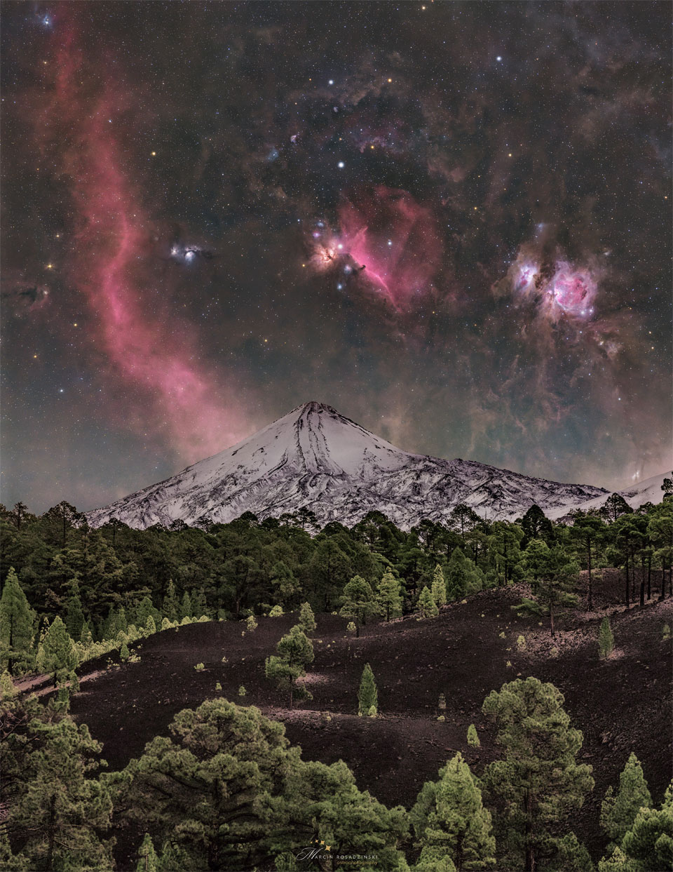 A starry sky is seen above a snowy volcano peak. The sky is
bright with many stars and red nebula. In the foreground is dark
soil and green evergreen trees. 
Więcej szczegółowych informacji w opisie poniżej.