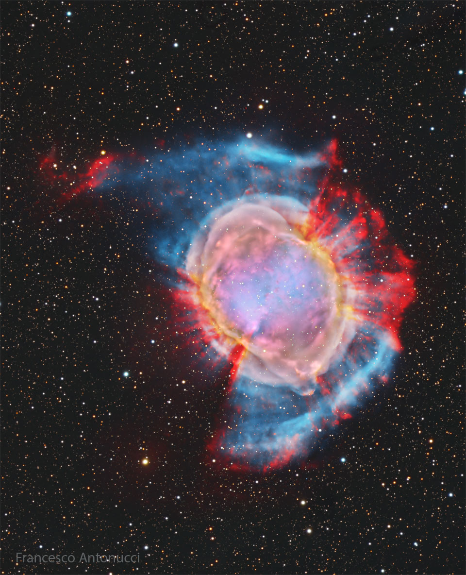 A starfield surrounds a large and colorful nebula.
Red spokes poke out from the outside of the nebula in
some directions, while a blue haze appears in other
directions. The nebula center is multicolored. 
Więcej szczegółowych informacji w opisie poniżej.