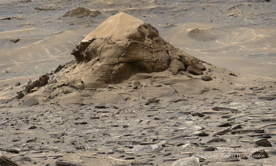 An orange landscape is seen with many darker rocks
visible in the foreground. Toward the image top is a large
rock outcrop with orange sand on top. The outcrop may appear
to resemble a large turtle.
Więcej szczegółowych informacji w opisie poniżej.