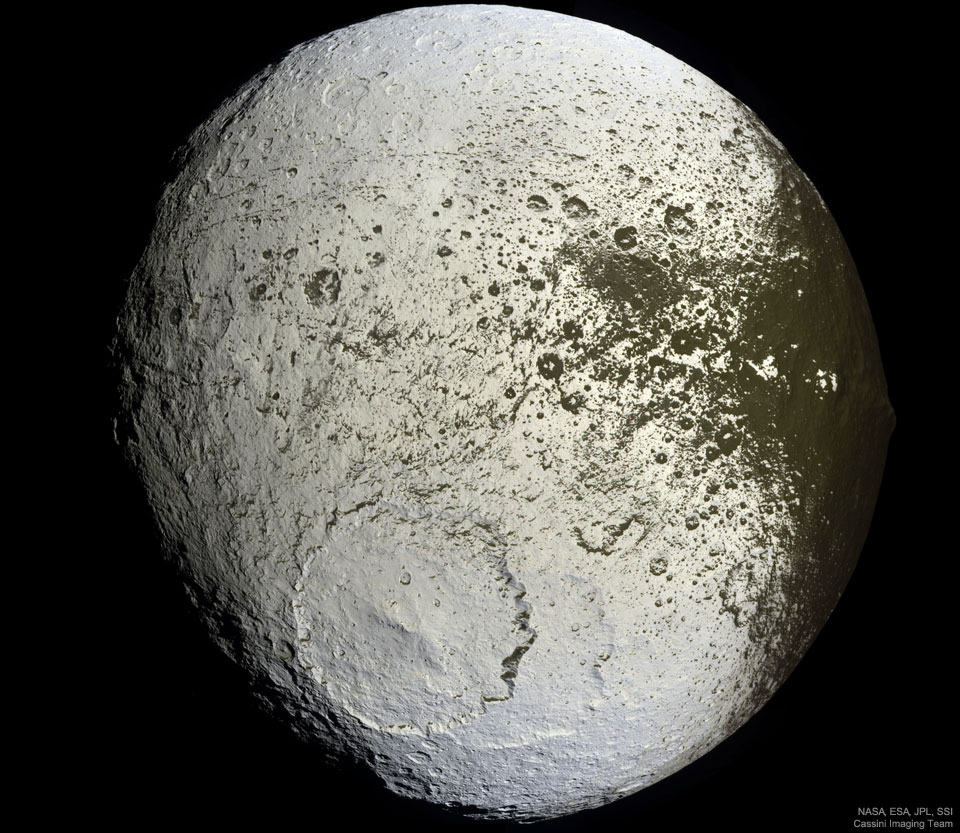 An unusually colored ball is shown against a 
dark background. The ball has numerous craters but
a very large cratered near the lower center. Much of
the ball is light colored, but around the right and
top it is green-brown, with a border area that seems
splattered. 
Więcej szczegółowych informacji w opisie poniżej.