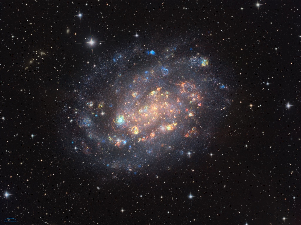 A bright spiral galaxy shows stars and colorful clous of gas
	  in red, green and blue colors. The background is a dark field of stars. 