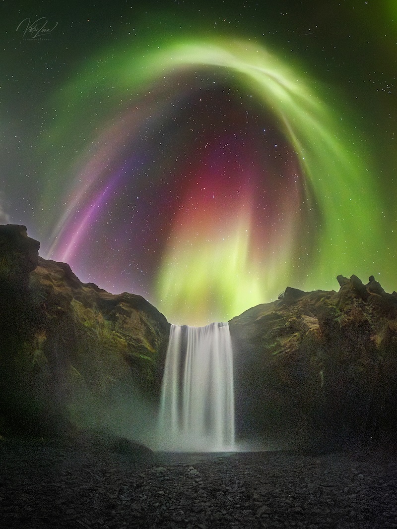 A night view of a waterfall with a colorful aurora in shades
	  of green, yellow, red and purple, and stars in the background.