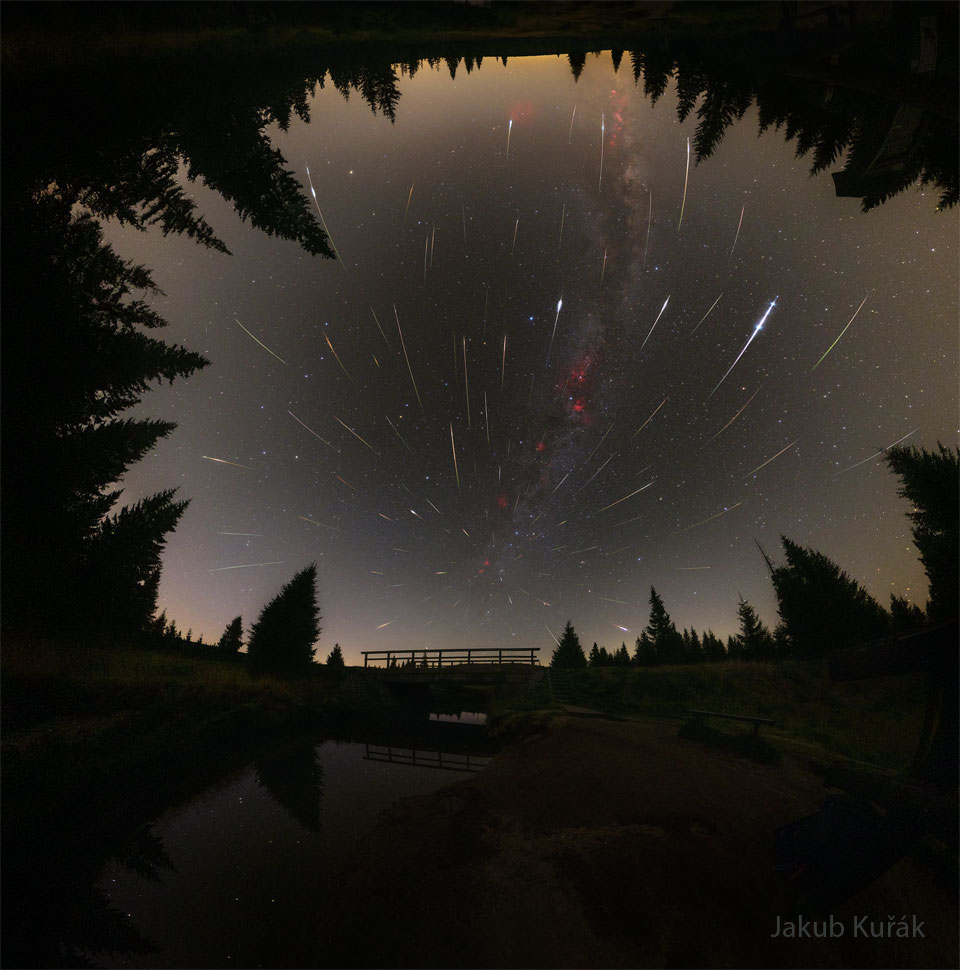A starfield is bounded by trees on three sides and
a pond on the bottom. Amongst the stars are many streaks
of light appearing to emanate from a place on the sky
just above a foreground bridge. 
Więcej szczegółowych informacji w opisie poniżej.