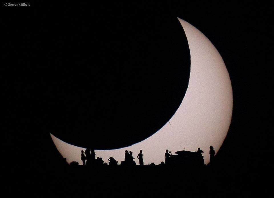 A light disk is visible on the lower left but the
rest of the disk is eclipsed by a darker disk of the 
same apparent size. At the bottom of the image are the
silhouettes of several people. 
Więcej szczegółowych informacji w opisie poniżej.