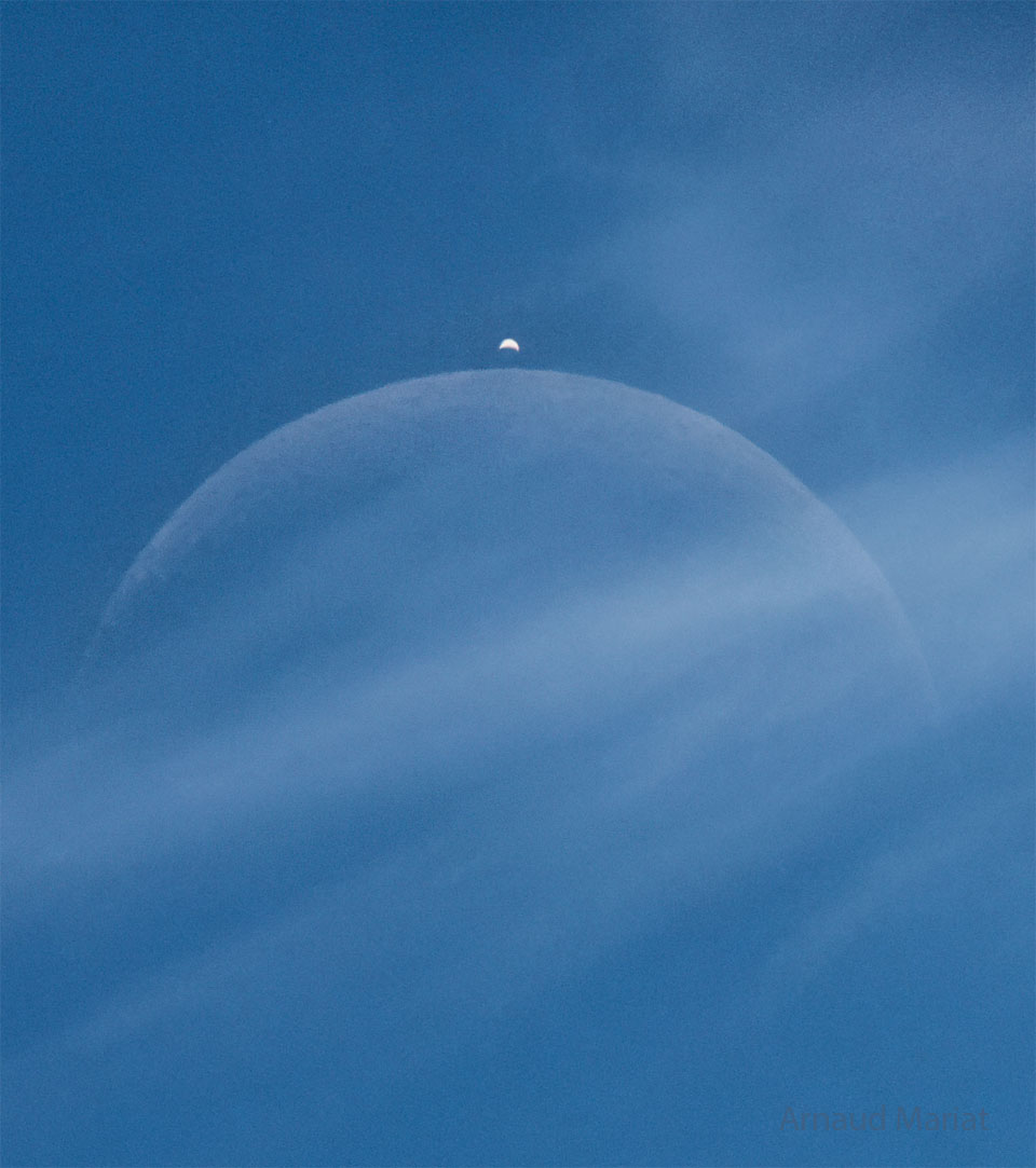 A blue sky has a light cloud crossing the middle of 
the image. Two crescent objects are visible through the 
cloud, a large faint crescent in the center and a brighter
crescent above it. 
Więcej szczegółowych informacji w opisie poniżej.