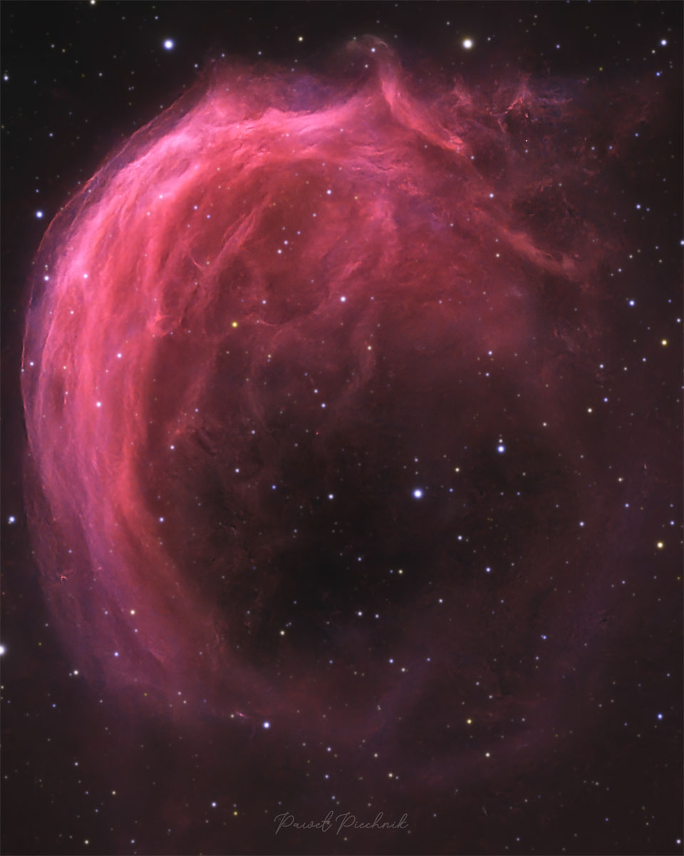 A starfield has a bright pink swirling nebula 
in it. The top part of the nebula shows much detail,
while the bottom part is mostly open.
Więcej szczegółowych informacji w opisie poniżej.